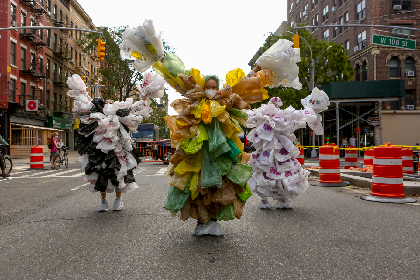 Down a New York street rimmed with traffic cones, wo women covered in white plastic bags walk behind a woman in green and yellow plastic bags. They all have their arms in a 5. 